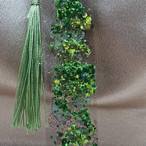 May include: A clear resin bookmark with green glitter and a green tassel. The glitter includes various shades of green and iridescent pieces. The tassel is made of green thread.