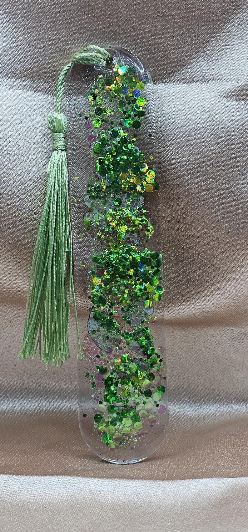 May include: A clear resin bookmark with green glitter and a green tassel. The glitter includes various shades of green and iridescent pieces. The tassel is made of green thread.