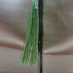 May include: A handmade bookmark with a dark, rectangular body and a light green tassel. The tassel is made of thin, twisted threads. The bookmark is vertical and appears to be made of resin or glass.