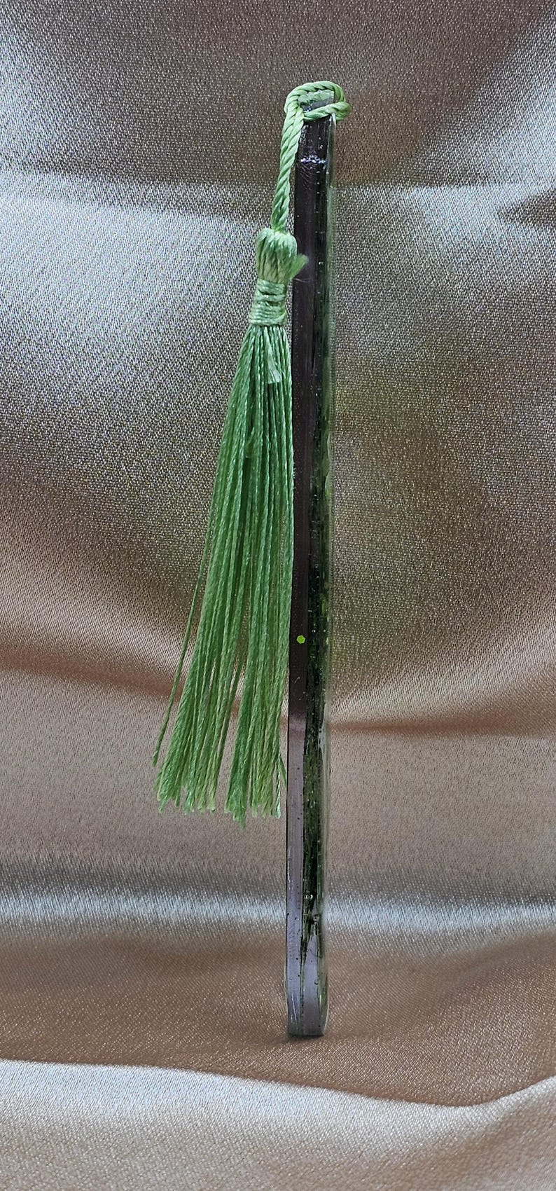 May include: A handmade bookmark with a dark, rectangular body and a light green tassel. The tassel is made of thin, twisted threads. The bookmark is vertical and appears to be made of resin or glass.