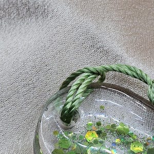 May include: Clear, oval-shaped resin pendant filled with green glitter and a green rope loop. The glitter is in various shades of green and shapes. The pendant is on a textured, light gray surface.