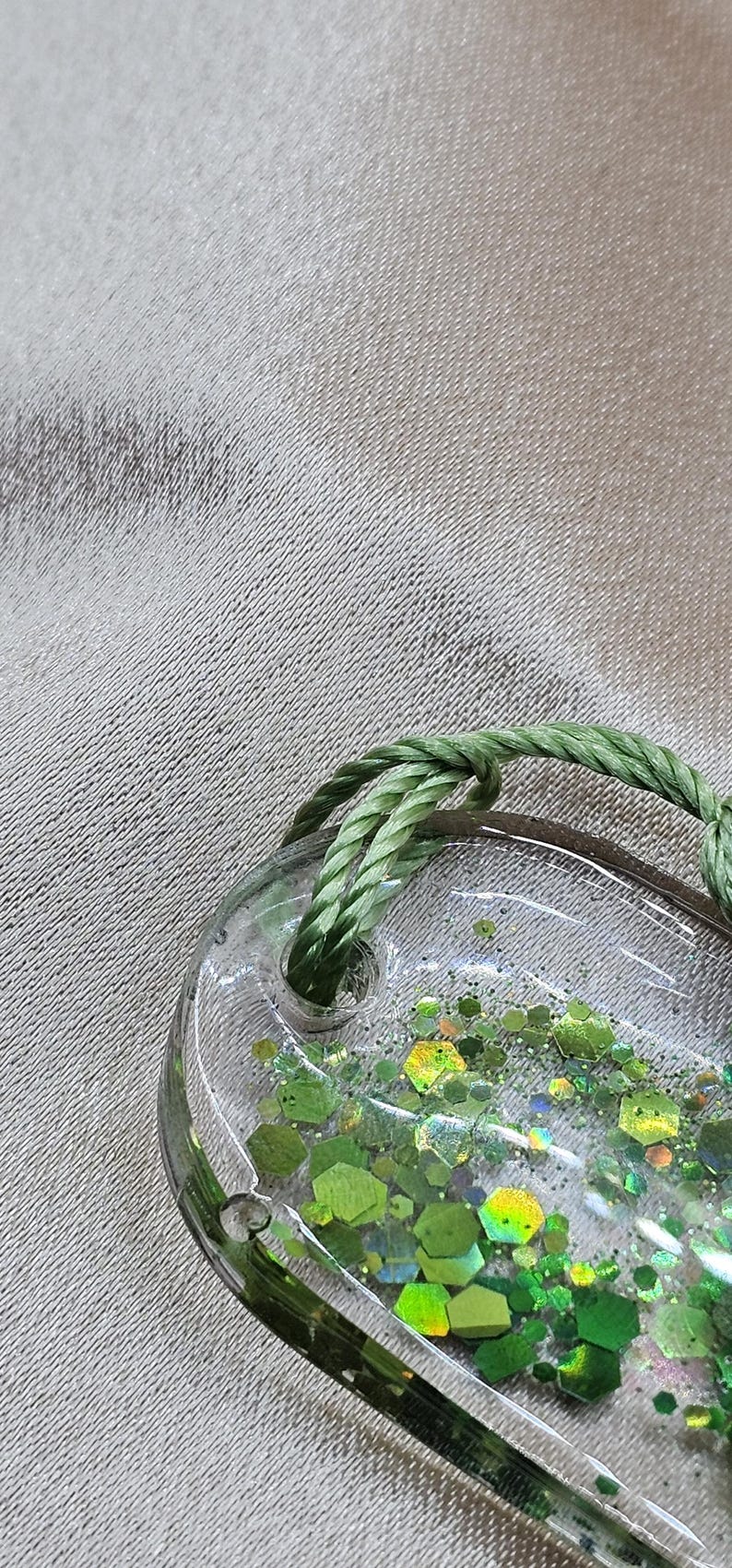 May include: Clear, oval-shaped resin pendant filled with green glitter and a green rope loop. The glitter is in various shades of green and shapes. The pendant is on a textured, light gray surface.