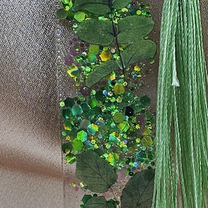 May include: A clear resin bookmark with embedded green leaves and glitter. The bookmark has a light green tassel attached to the top. The glitter is a mix of green, yellow, and iridescent colors.