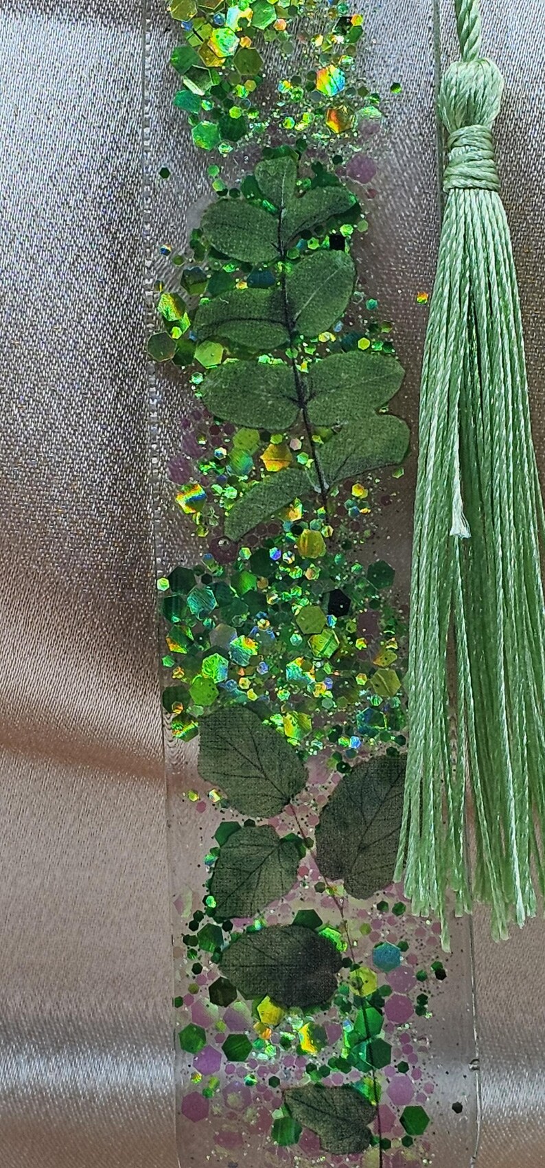 May include: A clear resin bookmark with embedded green leaves and glitter. The bookmark has a light green tassel attached to the top. The glitter is a mix of green, yellow, and iridescent colors.