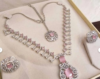 May include: A silver-toned jewelry set featuring a necklace, earrings, and a ring, all adorned with pink gemstones and clear crystals. The necklace has a pendant with a large pink stone. The set is displayed in a beige box.