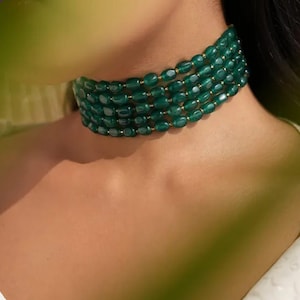 May include: A multi-strand choker necklace with rows of oval, dark green beads. Small gold-coloured spacers connect the beads. The necklace is worn around the neck, with the skin of the neck and shoulder visible.