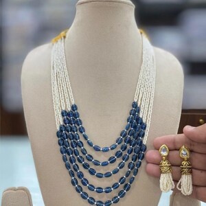 May include: A multi-strand necklace featuring navy blue oval beads and white seed beads. The necklace is displayed on a mannequin. Matching earrings with white tassels are held in the hand.