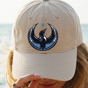 May include: A tan baseball cap featuring a circular emblem with a black bird design, a blue outline, and a star. The cap is worn by a person with long blonde hair, with the ocean in the background.