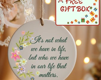 May include: A round, beige ceramic ornament hanging from a jute string. The ornament features a floral wreath design on the left side and the text: It's not what we have in life, but who we have in our life that matters. A gift box graphic is included.