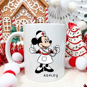 Peut inclure: Mug en c&eacute;ramique blanche avec un personnage de dessin anim&eacute; d'infirmi&egrave;re tenant un st&eacute;thoscope et faisant un signe de pouce lev&eacute;. Le personnage porte un uniforme d'infirmi&egrave;re. Le nom "ASHLEY" est &eacute;crit en script noir. D&eacute;corations de f&ecirc;te en arri&egrave;re-plan.