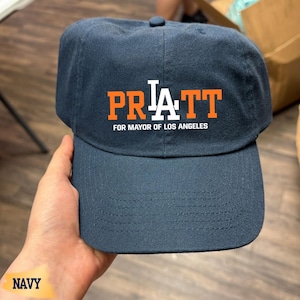 May include: A navy blue baseball cap featuring the orange text PRATT with a white LA logo integrated into the center. Below the name, the text FOR MAYOR OF LOS ANGELES is printed in white. The cap has a structured crown and a curved brim.