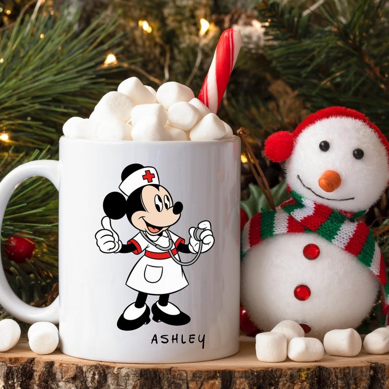 Peut inclure: Mug en c&eacute;ramique blanche avec un dessin anim&eacute; de Minnie Mouse en infirmi&egrave;re, avec le nom "ASHLEY" en dessous. La tasse est remplie de guimauves et d'un sucre d'orge. Une figurine de bonhomme de neige est &agrave; c&ocirc;t&eacute; de la tasse, avec une &eacute;charpe verte et rouge.