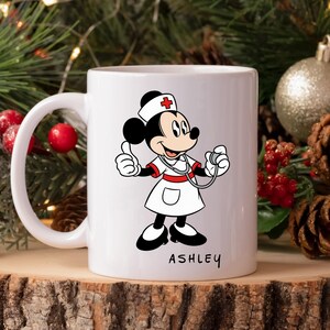 Peut inclure: Mug en c&eacute;ramique blanche avec un dessin anim&eacute; de souris habill&eacute;e en infirmi&egrave;re, portant un uniforme blanc avec une croix rouge sur le chapeau. La souris tient un st&eacute;thoscope et fait un signe de pouce lev&eacute;. Le nom "ASHLEY" est imprim&eacute; sur le mug.