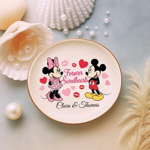 May include: A round, cream-colored plate featuring Mickey and Minnie Mouse. The plate has a brown rim and includes the text "Forever Sweethearts" with hearts and lip graphics. Personalized with the names "Clara & Thomas".