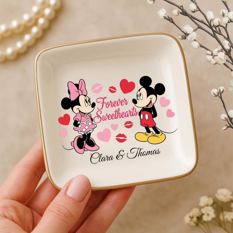May include: A square, off-white trinket dish with a gold rim. The dish features Minnie and Mickey Mouse, hearts, and the words "Forever Sweethearts" and "Clara & Thomas". The characters are in color, with Minnie in pink and Mickey in red and yellow.