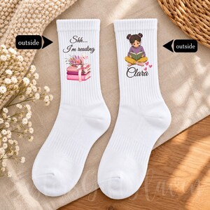 May include: A pair of white crew socks. The left sock features a stack of pink books with a bow, flowers, and the text "Shh... I'm reading". The right sock displays an illustration of a person reading a book with the name "Clara" printed below.