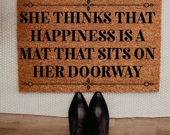 May include: A rectangular doormat with the text "SHE THINKS THAT HAPPINESS IS A MAT THAT SITS ON HER DOORWAY" in black. The mat is brown with decorative black borders. Black boots and blue jeans are visible.