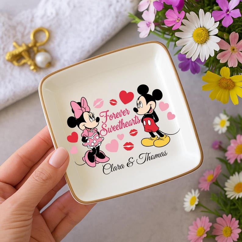 May include: A square, white ceramic trinket dish with a gold rim. The dish features a graphic of Mickey and Minnie Mouse, hearts, and the words "Forever Sweethearts." The names "Clara & Thomas" are also printed on the dish.