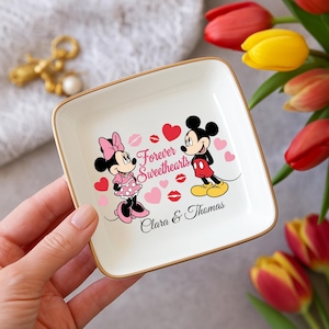 May include: A square, white ceramic trinket dish with a gold rim. The dish features a graphic of Mickey and Minnie Mouse, hearts, and the words "Forever Sweethearts." The names "Clara & Thomas" are also printed on the dish.