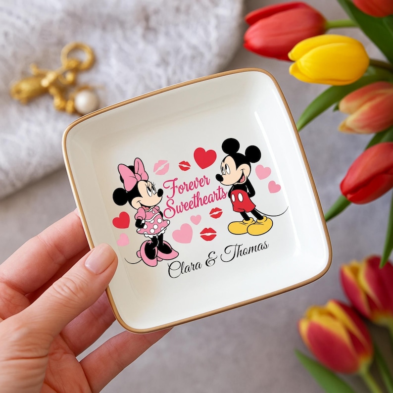 May include: A square, white ceramic trinket dish with a gold rim. The dish features a graphic of Mickey and Minnie Mouse, hearts, and the words "Forever Sweethearts." The names "Clara & Thomas" are also printed on the dish.