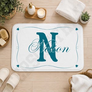 May include: A rectangular white bath mat featuring a large teal letter N in the center with the name Nelson written in a cursive teal script across it. The mat has a thin teal border with decorative flourishes at the corners and sides.
