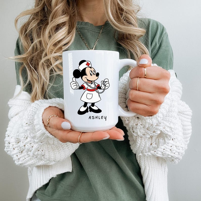 Peut inclure: Mug en c&eacute;ramique blanche avec Minnie Mouse en infirmi&egrave;re, avec un st&eacute;thoscope et une coiffe d'infirmi&egrave;re. Le mug porte le nom "ASHLEY" en noir. Le design comprend des accents rouges et noirs.