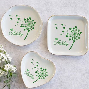 May include: Three white ceramic dishes with gold rims, in round, square, and heart shapes. Each features a green dandelion design with the name "Ashley" in cursive. The dandelion seeds are blowing away, and a green heart is next to the name.