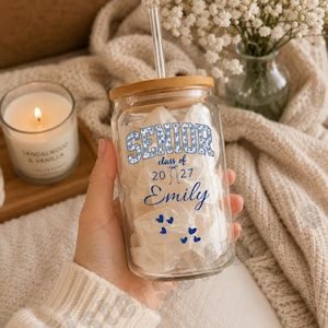 May include: A clear glass tumbler with a bamboo lid and a clear straw. The glass features the blue text SENIOR, class of 2027, and the name Emily in blue script. Small blue heart icons are printed below the name, and the glass is filled with white paper.