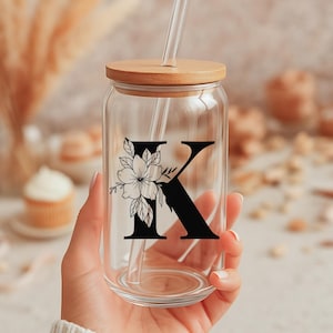 Peut inclure: Un gobelet en verre transparent en forme de canette avec un couvercle en bois et une paille. La lettre "K" est affichée en noir avec un motif floral. Le gobelet est tenu dans une main.