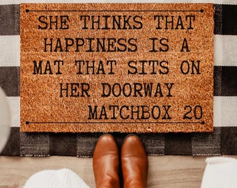 May include: A rectangular doormat with the text "SHE THINKS THAT HAPPINESS IS A MAT THAT SITS ON HER DOORWAY MATCHBOX 20" in black letters. The mat is brown and sits on a black and white checkered rug. Brown boots are visible.