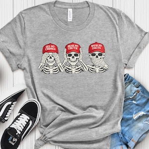 May include: A gray t-shirt featuring a graphic of three skeletons in red caps, each in the classic "see no evil, hear no evil, speak no evil" pose. The caps read "See No Crimes," "Hear No Truth," and "Speak No Facts."