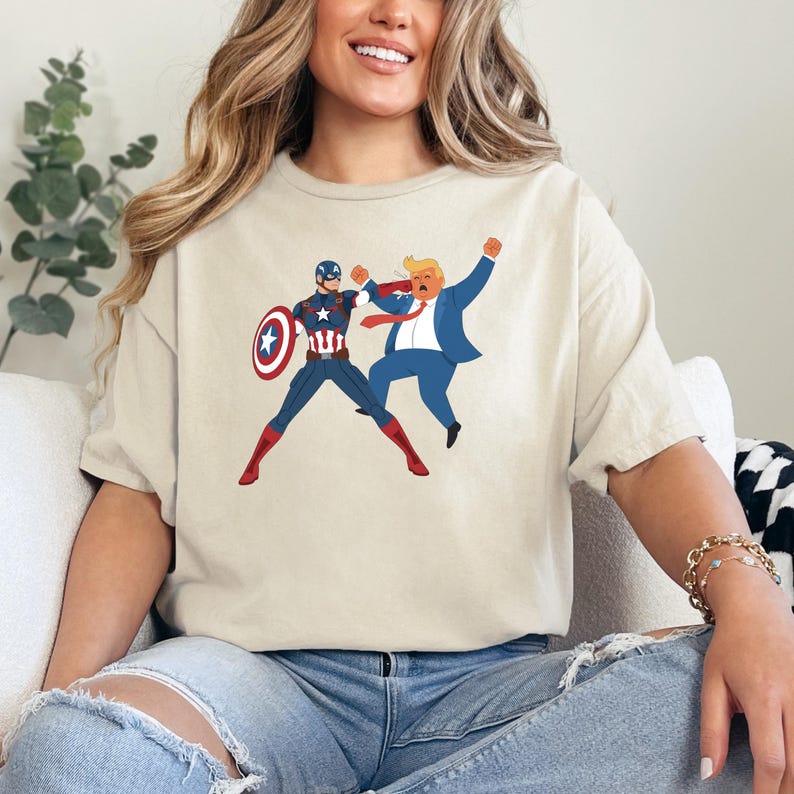 May include: Beige t-shirt featuring a cartoon illustration of Captain America and a man in a blue suit in a fighting pose. Captain America is in a blue and red suit with a shield. The other man is wearing a red tie.