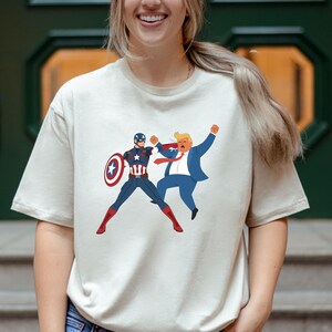 May include: A cream-coloured t-shirt with a cartoon graphic of Captain America in a fight with a cartoon version of Donald Trump. Captain America is in a blue and red suit with a shield. Trump is in a blue suit and red tie.