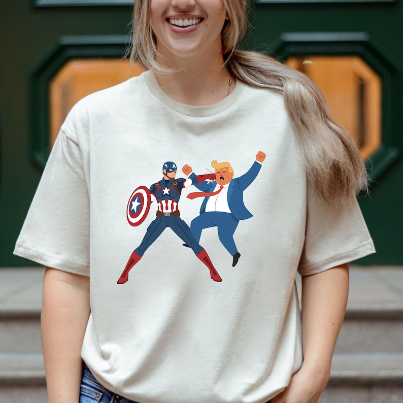 May include: A cream-coloured t-shirt with a cartoon graphic of Captain America in a fight with a cartoon version of Donald Trump. Captain America is in a blue and red suit with a shield. Trump is in a blue suit and red tie.