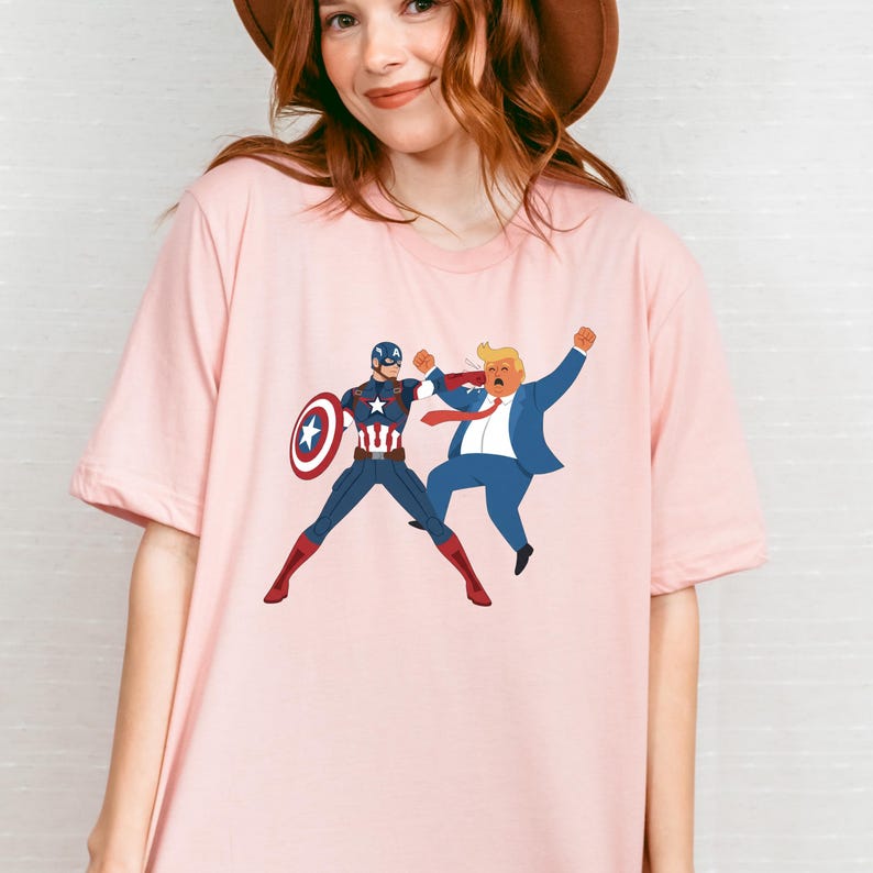 May include: Pale pink t-shirt with a cartoon graphic of Captain America punching a figure in a blue suit. Captain America is in a blue and red suit with a shield. The other figure has blonde hair and a red tie.