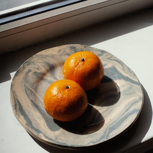 May include: Two bright orange tangerines rest on a decorative plate with a marbled blue and beige pattern. The plate sits on a white surface near a window, with sunlight casting shadows.
