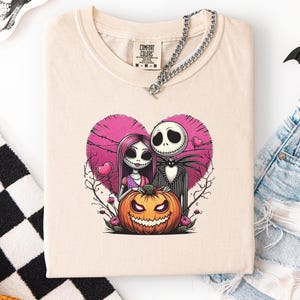 May include: Cream-colored t-shirt featuring a pink heart design with Jack Skellington and Sally from The Nightmare Before Christmas. A carved orange pumpkin sits below them. The shirt has a Comfort Colors tag and a silver chain necklace. Black and white checkered fabric and a skull are visible.