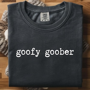 May include: Dark gray t-shirt with the words "goofy goober" in white, displayed on a wooden surface. The shirt is folded and rests on a textured, light brown fabric. The shirt has a crew neck and a tag.