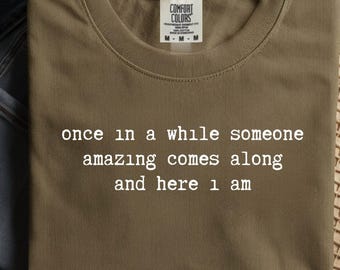 May include: Olive green t-shirt with the text "once in a while someone amazing comes along and here 1 am" in white. The shirt has a crew neck and short sleeves. The tag reads "Comfort Colors".