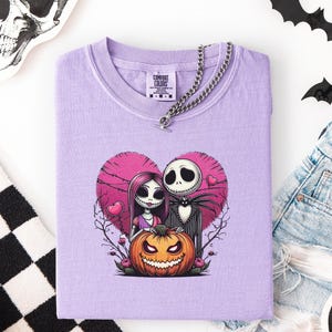 May include: A lilac-colored t-shirt featuring a graphic of Jack and Sally from The Nightmare Before Christmas, set against a pink heart and a jack-o'-lantern. The shirt has a chain necklace and the Comfort Colors brand label.