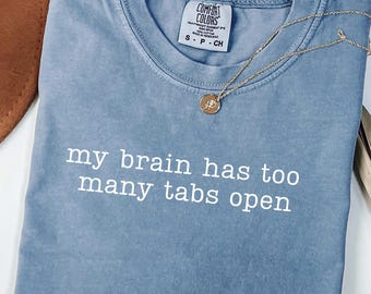 May include: A blue Comfort Colors t-shirt with the text "my brain has too many tabs open" in white. A gold necklace with a pendant is draped across the shirt. A brown hat is visible in the upper left corner.