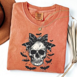 K&ouml;nnte beinhalten: Orangefarbenes Comfort Colors T-Shirt mit einem Totenkopf-Motiv, einer schwarz-wei&szlig;en Schleife und Flederm&auml;usen. Das T-Shirt mit Rundhalsausschnitt hat kurze &Auml;rmel. Eine goldene Halskette mit Anh&auml;nger ist sichtbar. Das T-Shirt ist Gr&ouml;&szlig;e S.
