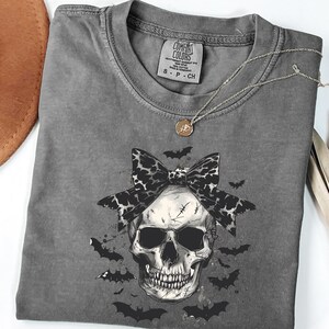 May include: A gray Comfort Colors t-shirt with a skull graphic. The skull has a black and white bow and is surrounded by bats. A silver necklace with a small pendant is draped on the shirt. The shirt has the Comfort Colors logo on the neckline.