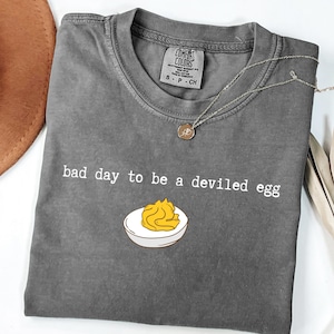 May include: Gray Comfort Colors t-shirt with the phrase "bad day to be a deviled egg" in white text. A cartoon illustration of a deviled egg is below the text. A silver necklace with a small pendant is on the shirt.