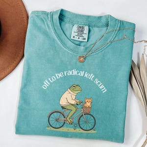 May include: Teal Comfort Colors t-shirt with the phrase "off to be radical left scum" in a curved font. The graphic shows a frog in a sweater riding a bicycle with a cat in the basket. A gold necklace and a brown hat are also visible.