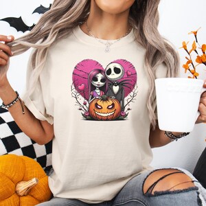 May include: Beige t-shirt featuring a Halloween-themed graphic. The design includes Jack Skellington and Sally from The Nightmare Before Christmas, a pink heart, and a carved pumpkin. The shirt is a casual, crew neck style.