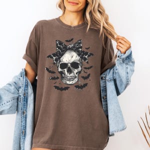K&ouml;nnte beinhalten: Braunes T-Shirt mit einem Totenkopf-Motiv, einer schwarz-wei&szlig; gemusterten Schleife und Fledermaus-Silhouetten. Der Totenkopf ist detailliert schattiert, und die Flederm&auml;use erzeugen ein gruseliges Halloween-Design.