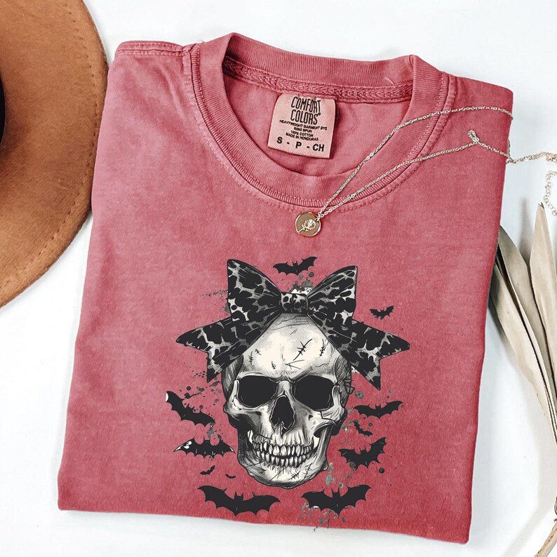 K&ouml;nnte beinhalten: Ein rotes Comfort Colors T-Shirt mit einem Totenkopf-Motiv, einer Leopardenmuster-Schleife und Flederm&auml;usen. Eine silberne Halskette mit Anh&auml;nger liegt &uuml;ber dem Shirt. Das Shirt ist in Gr&ouml;&szlig;e S.