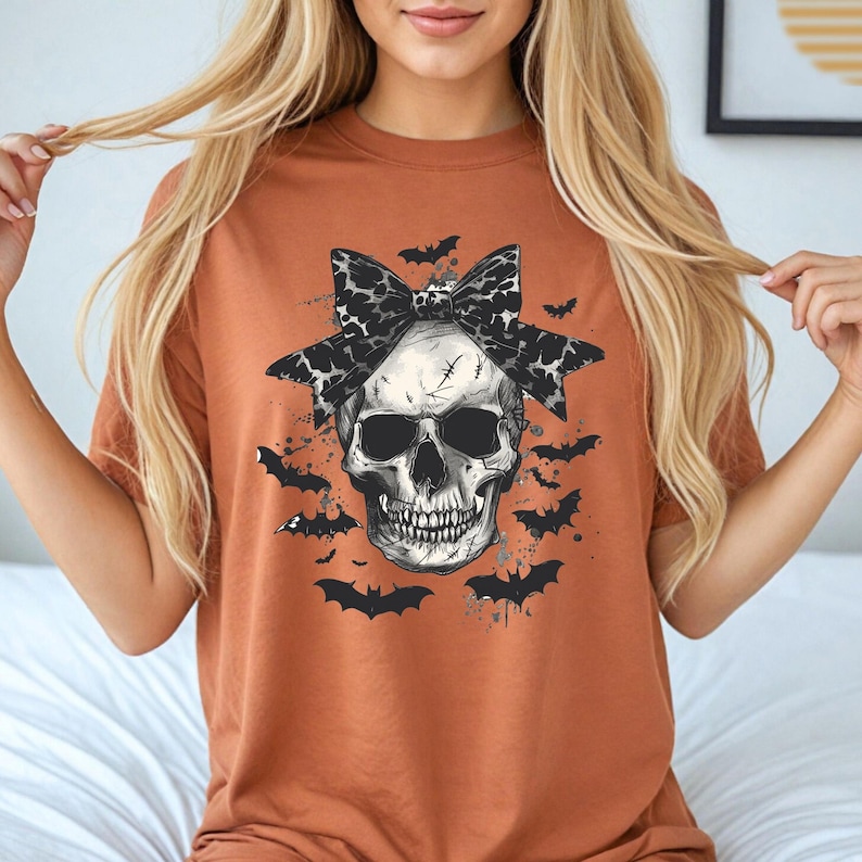 K&ouml;nnte beinhalten: Rostfarbenes T-Shirt mit detaillierter Totenkopf-Grafik, Leopardenmuster-Schleife und Flederm&auml;usen. Der Sch&auml;del hat sichtbare Z&auml;hne und Augenh&ouml;hlen. Das Design ist von mehreren Fledermaus-Silhouetten umgeben, was ein Halloween-Thema erzeugt.