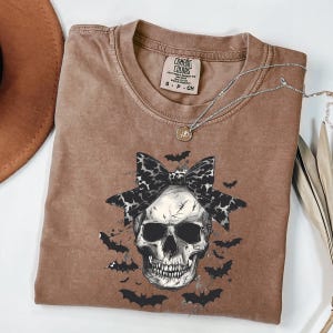 K&ouml;nnte beinhalten: Ein braunes Comfort Colors T-Shirt mit einem Totenkopf-Motiv, einer Leopardenmuster-Schleife und Flederm&auml;usen. Das T-Shirt mit Rundhalsausschnitt hat kurze &Auml;rmel. Eine silberne Halskette mit einem kleinen Anh&auml;nger liegt &uuml;ber dem Shirt.